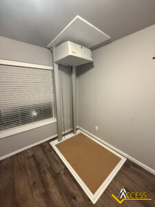 A residential wheelchair lift with a platform and vertical tracks installed in a corner of a room with gray walls, wood flooring, and a window with blinds. The Access logo appears in the bottom right corner.