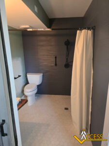 Accessible bathroom with a white toilet, walk-in shower with a black showerhead, grab bar, and cream shower curtain. The floor has white hexagonal tiles and there is the Access logo in the bottom right corner.