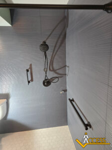 Accessible shower with dark gray textured wall, two black grab bars, a handheld showerhead, and white hexagonal floor tiles; Access logo is visible in the bottom right corner.