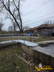 A metal wheelchair ramp leads from a concrete platform down to a grassy yard with fallen leaves. Leafless trees and residential houses are visible in the background. The Access company logo is in the bottom right corner.