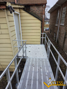 A metal wheelchair ramp with railings leads to a door on a yellow house. The ramp is surrounded by brick buildings, and the ACCESS logo is in the bottom right corner.