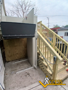 A vertical platform lift is installed next to a wooden staircase outside a building, providing wheelchair accessibility. The lift is positioned on a concrete surface, and the stairs have wooden railings. Access company logo is visible.
