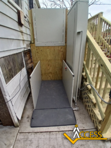 An outdoor vertical platform lift next to a wooden ramp and stairs, providing wheelchair access to a raised entryway. The Access company logo is visible in the lower right corner.