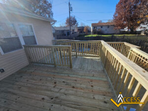 A newly built wooden deck with railings attached to a beige house, overlooking a backyard with grass, trees, and neighboring houses; the Access company logo is in the bottom right corner.