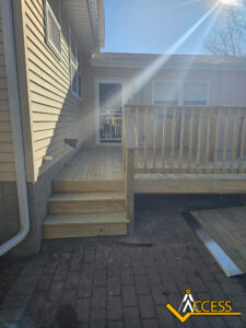 A wooden deck with steps leading to a door, attached to a beige house. Sunlight shines across the deck, and a brick patio is below. A ramp is partially visible on the right. The Access company logo is in the bottom right.