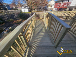 A wooden wheelchair ramp with railings leads to a backyard deck on a sunny day. The yard has a fence, trees, and some landscaping. The Access company logo appears in the lower right corner.