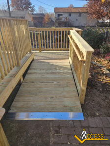 A wooden wheelchair ramp with safety railings on both sides leads from a brick path to a raised deck. The ACCESS company logo is visible in the bottom right corner. Houses and autumn trees are in the background.