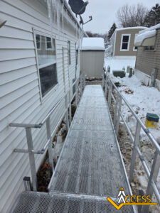 A metal wheelchair ramp with railings is attached to a house in a snowy yard. Icicles hang from the roof, and a shed and mobile homes are visible in the background. The ACCESS logo is in the lower right corner.