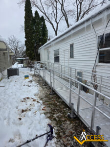 A metal wheelchair ramp is installed alongside a white house in a snowy yard. Trees and a shed are visible in the background. The ground is partially covered with snow and a snow shovel lies nearby. Access company logo is in the corner.