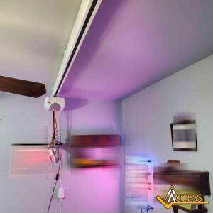 A white motorized curtain track is mounted on the ceiling near a wall, with attached wires and a remote control. The room has blurred colorful lights and paintings on the wall. Access logo appears in the lower right corner.
