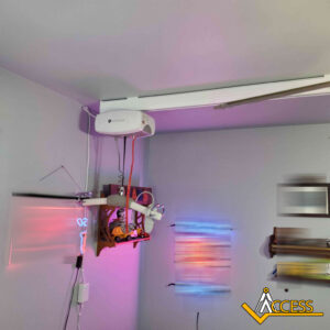 Ceiling-mounted track lift system with a white motor unit and red straps attached to a wooden harness, in a room with blurred colorful lights and an Access logo in the corner.
