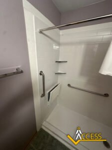 Accessible shower with white tiles, grab bars, a folding shower seat, wall-mounted corner shelves, and a curtain rod. ACCESS logo is visible in the bottom right corner.