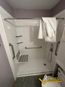 Accessible shower with a fold-down seat, grab bars, handheld showerhead, corner shelves, white tile walls, and a partially open shower curtain. The Access logo is visible in the bottom right corner.