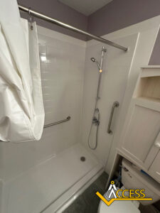 Accessible shower with white tiles, a handheld showerhead, grab bars on two walls, a white curtain partly open, and a cabinet above a toilet. “Access” logo is in the bottom right corner.