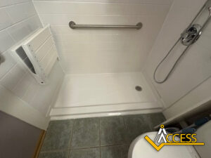 Accessible shower with a fold-down seat, grab bar, handheld showerhead, and tiled floor. The Access logo is visible in the bottom right corner.