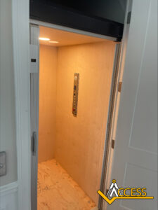 A small residential elevator with wood-paneled walls and floor, a control panel on the back wall, and the Access company logo in the lower right corner. The elevator door is partially open.
