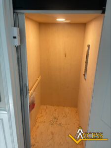 A small residential elevator with unfinished wooden walls and floor, a handrail on the left, and control panel on the right. The elevator is empty and illuminated by a ceiling light. ACCESS logo is in the bottom right corner.