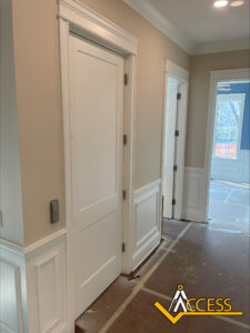 A hallway with beige walls, white trim, and wainscoting features two closed white doors and one open door. The floor is covered for protection, and sunlight streams through windows. An “ACCESS” logo is in the bottom right corner.