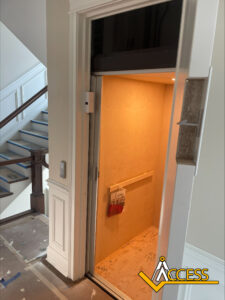 A small residential elevator with wooden interior and handrail is open next to a staircase. The elevator has some papers on the floor. The Access logo is visible in the bottom right corner.