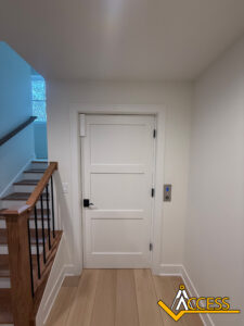 A white residential elevator door next to a staircase with wooden handrails, light wood floor, and an elevator control panel on the wall. The Access company logo is in the bottom right corner.