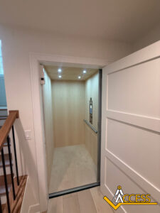 A modern home elevator with wood-paneled interior and recessed ceiling lights is seen with its white sliding door open. A staircase and ACCESS logo are visible in the foreground.
