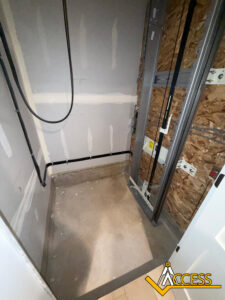 An unfinished shower area with exposed wall framing, plumbing pipes, and partially installed drywall. The floor is bare concrete and the walls have visible seams and construction materials. The word ACCESS is on the lower right.