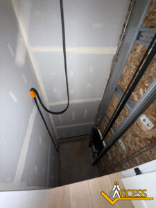 View looking down an unfinished residential elevator shaft with exposed drywall and plywood, visible cables, tracks, and Access logo in the bottom right corner.