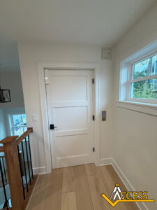 A white door with a black handle is at the top of a stairway next to a window. There is an elevator call button on the wall beside the door. Hardwood floors and a railing are visible. The Access logo is in the bottom right corner.