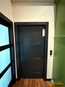 A modern black door with three rectangular panels, set in a white wall with wood flooring. To the right of the door is a keypad or touchpad, and the Access logo is visible in the lower right corner.