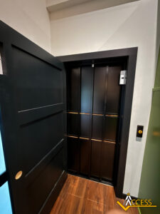 A small, modern residential elevator with black sliding doors and gold accents, located in a hallway with wood flooring and white walls. A call button is mounted on the right wall.