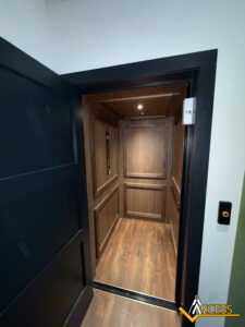 A modern residential elevator with wood-paneled interior and flooring, seen through an open black door. Elevator controls are on the left wall; a small ceiling light illuminates the space. Access logo is visible in the bottom right corner.
