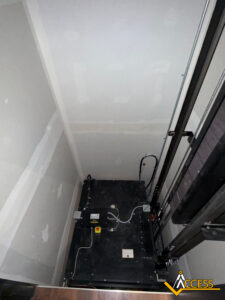 View from above of an empty elevator shaft with a partially installed elevator platform, visible cables, control boxes, and bare walls. The Access company logo is in the bottom right corner.