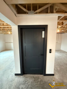A black door with a rectangular panel design is set in a white wall inside a room with unfinished floors and an exposed wooden ceiling. A black control panel is mounted on the wall next to the door. Access logo in bottom right corner.