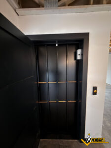 A modern black elevator door with vertical gold accents is set in a white wall. A round elevator call button is mounted beside the door, and a logo reading Access appears in the lower right corner.