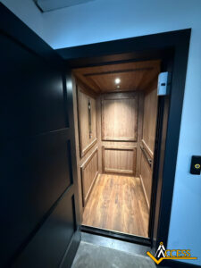 An open modern home elevator with wood-paneled walls and flooring, softly lit from above. The elevator is viewed from outside the open black door, and the Access company logo is visible in the bottom right corner.