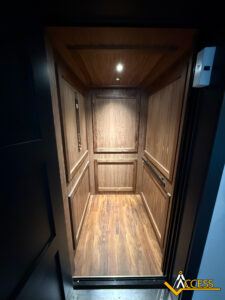A small, wood-paneled residential elevator with a single overhead light, open door, and control panel on the left wall. The floor and walls are finished in matching dark wood. Access logo is visible in the corner.