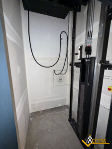 An unfinished elevator shaft with exposed wiring, metal rails, and concrete floor. The walls are unpainted drywall, and there is an Access company logo in the bottom right corner.