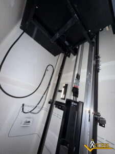 A view inside an elevator shaft showing cables, rails, and mechanical components. The walls are unfinished, with visible wiring. The Access logo is in the lower right corner.