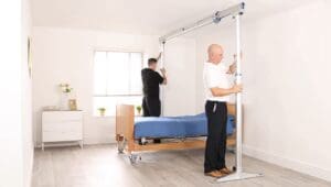 Two men are assembling a Mackworth FSG 440 Gantry over a hospital bed in a bright, tidy room featuring wooden flooring, a window, and a white dresser decorated with flowers and a photo frame.