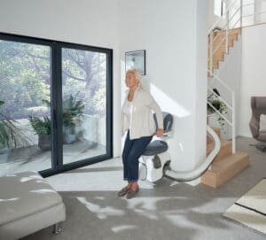 An older woman stands up from a Windy City Stair Lifts chair at the base of a modern staircase in a bright, sunlit living room with large windows, indoor plants, and contemporary furniture.