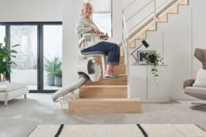 An older woman smiles while using a stair lift by Access Elevator to go up the stairs in a modern, bright living room, featuring plants and contemporary furniture.