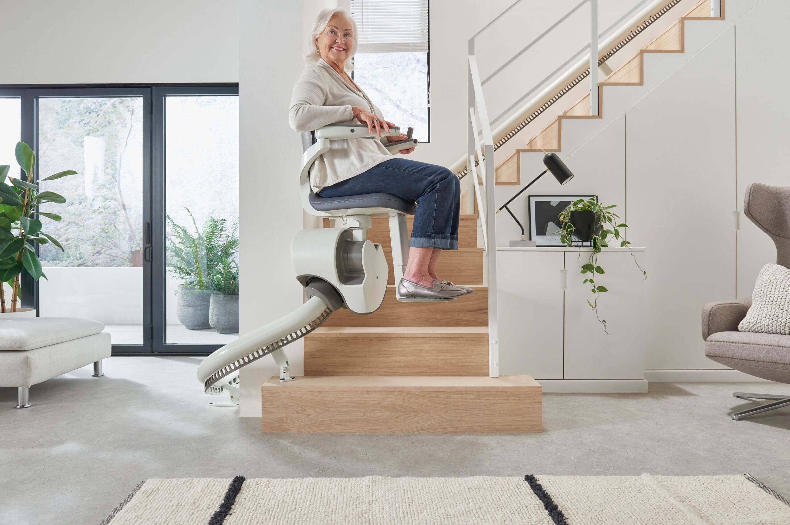 An older woman smiles while using a stair lift by Access Elevator to go up the stairs in a modern, bright living room, featuring plants and contemporary furniture.