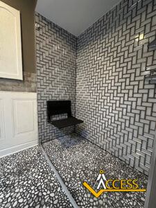 A modern accessible shower features a wall-mounted black seat, geometric patterned tiles on the walls, pebble-style floor tiles, and the ACCESS logo at the bottom right corner.