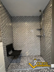 A modern shower with a black fold-down seat, geometric tiled walls, a handheld showerhead, and pebble-style floor tiles. The Access logo appears in the bottom right corner.