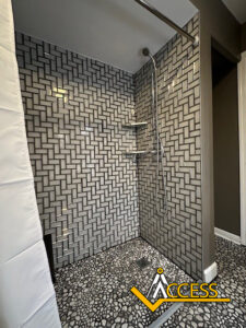 A modern shower with gray and white geometric wall tiles, pebble-patterned floor tiles, a handheld showerhead, built-in shelves, and the ACCESS logo in the bottom right corner.