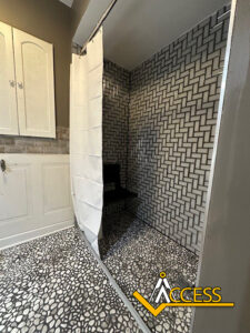 Modern accessible shower with a white curtain, black bench, and black-and-white geometric tile walls; pebble-patterned floor. White cabinets and gray wall outside the shower. ACCESS logo in bottom right corner.