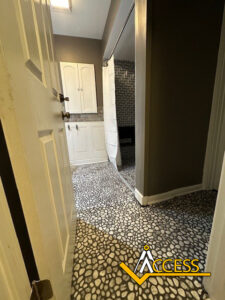 View from an open door into a bathroom with pebble-patterned floor, white cabinets, a shower with a white curtain, and dark gray walls. The ACCESS logo is at the bottom right corner.