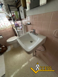 A small bathroom with pink tiled walls, a white wall-mounted sink, a toilet, shelves with towels and toiletries, and a window with floral curtains. The word ACCESS with a logo is at the bottom right corner.