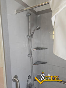 Shower with a handheld showerhead, grab bar, two corner shelves, chrome fixtures, and a white curtain partially closed; ACCESS logo appears in the lower right corner.