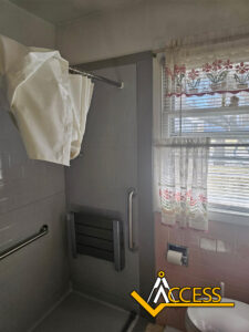 A bathroom with a walk-in shower, fold-down shower seat, grab bars, white shower curtain, and a window with sheer curtains. The ACCESS logo is in the lower right corner.